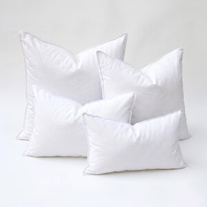 May include: Four white pillows of varying sizes are arranged on a white surface. The pillows have a soft, puffy appearance with a subtle texture. Each pillow has a white border, and the overall aesthetic is clean and minimalist.