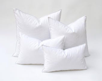 May include: Four white pillows of varying sizes are arranged on a white surface. The pillows have a soft, puffy appearance with a subtle texture. Each pillow has a white border, and the overall aesthetic is clean and minimalist.