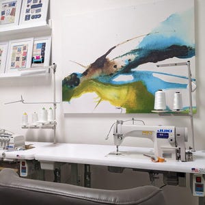 K&ouml;nnte beinhalten: Ein N&auml;hstudio mit einer wei&szlig;en Juki-N&auml;hmaschine auf einem gro&szlig;en wei&szlig;en Tisch. Der Raum umfasst Garnrollen, gerahmte Kunstwerke und einen grauen Lederstuhl. An der Wand h&auml;ngt ein abstraktes Gem&auml;lde mit blauen, gr&uuml;nen und braunen Farbt&ouml;nen.
