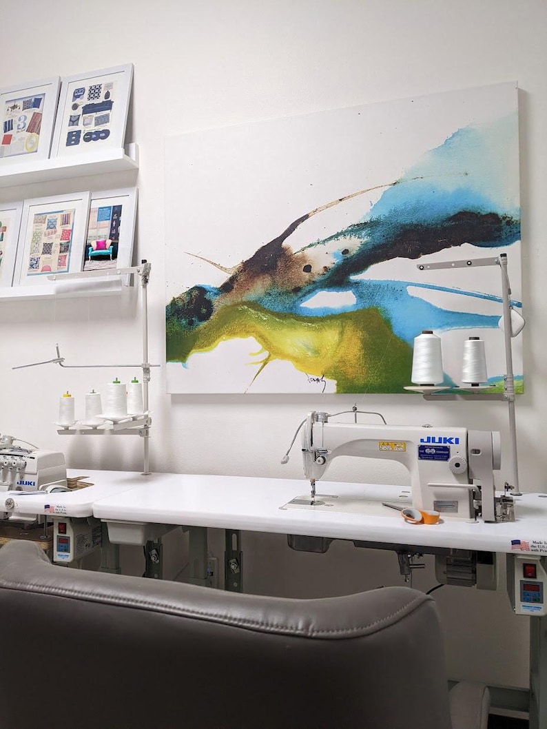 K&ouml;nnte beinhalten: Ein N&auml;hstudio mit einer wei&szlig;en Juki-N&auml;hmaschine auf einem gro&szlig;en wei&szlig;en Tisch. Der Raum umfasst Garnrollen, gerahmte Kunstwerke und einen grauen Lederstuhl. An der Wand h&auml;ngt ein abstraktes Gem&auml;lde mit blauen, gr&uuml;nen und braunen Farbt&ouml;nen.