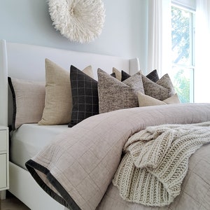 May include: A white bed with a light gray duvet cover and a white knitted throw blanket. The bed has a white headboard and several pillows in various shades of brown and gray. A white feather wall decor hangs above the bed.