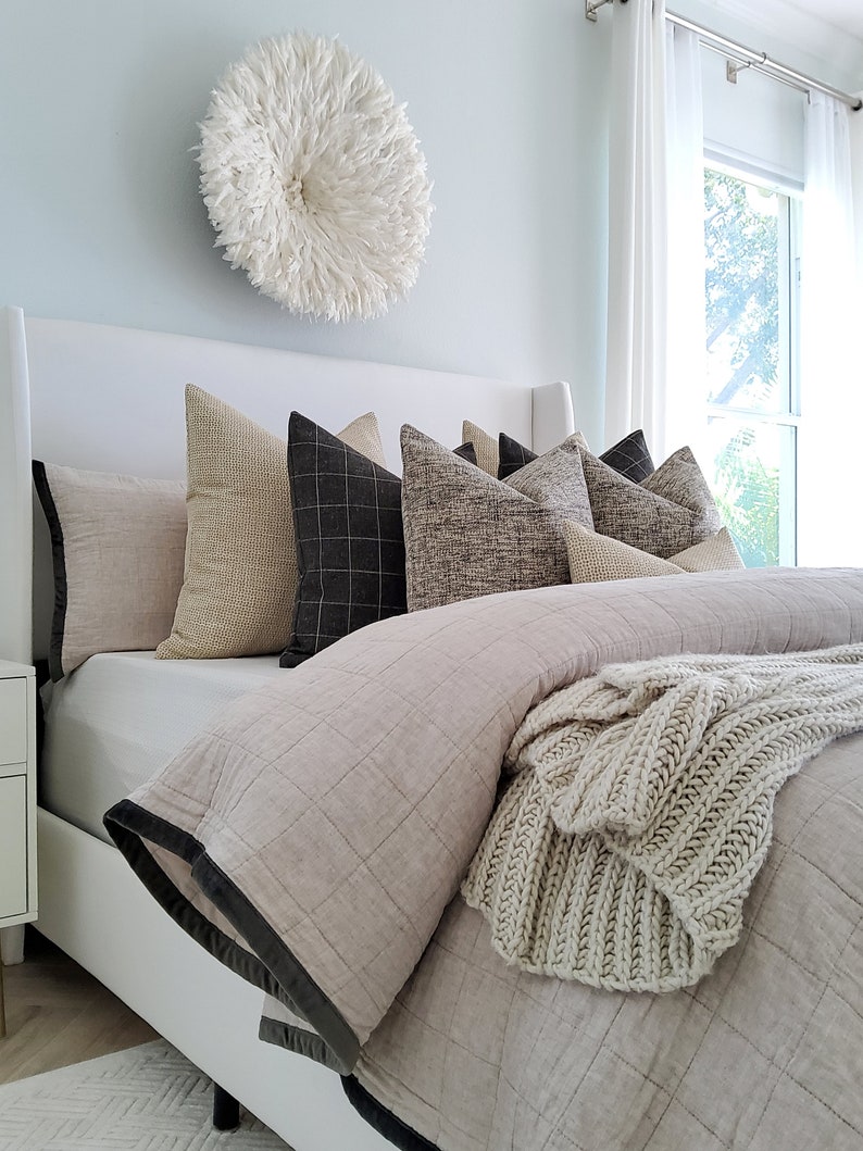 May include: A white bed with a light gray duvet cover and a white knitted throw blanket. The bed has a white headboard and several pillows in various shades of brown and gray. A white feather wall decor hangs above the bed.