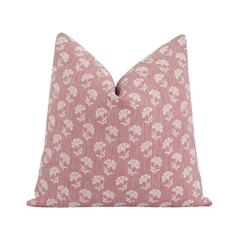 May include: A decorative throw pillow with a dusty rose background and a repeating pattern of white floral designs. The pillow has a textured appearance and a soft, inviting look, perfect for adding a touch of elegance to any room.