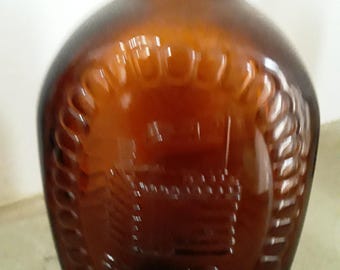 May include: A brown glass bottle with a rounded body and a narrow neck. The bottle has an embossed design featuring a building within a wreath. The glass has a dark amber color.