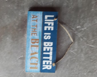 May include: A rectangular wooden wall sign featuring a two-tone design with a light blue textured section and a dark blue section. The sign displays the white and tan text "LIFE IS BETTER AT THE BEACH" and includes a tan rope for hanging.