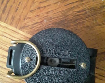 May include: A black Engineer directional compass with a textured surface. The compass has a gold-colored ring and hinge. The text "ENGINEER" and "DIRECTIONAL COMPASS" are printed on the compass.