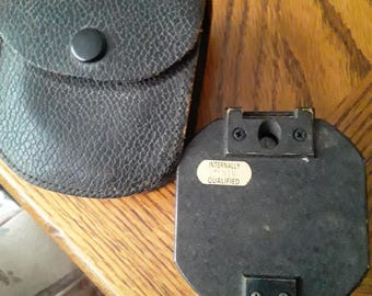 May include: A black leather case with a snap closure next to a black, rectangular device. The device has two screws and a sticker that reads "INTERNALLY NSN QUALIFIED". The items are on a wooden surface.
