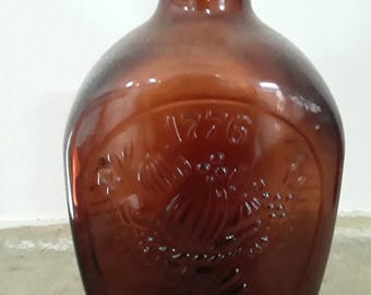 May include: A brown glass bottle with a rounded body and a narrow neck. The bottle has an embossed design featuring the number "1776" and an emblem. The bottle is empty and has a cork stopper.