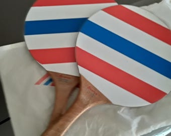 May include: Two ping pong paddles with red, white, and blue striped faces. The wooden handles are light brown. The paddles are resting on a white surface with a logo.