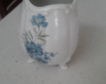 May include: A white ceramic vase with a blue floral design and three legs. The vase has a scalloped rim and a slightly rounded shape.