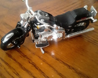 May include: A black and silver toy motorcycle with a black seat and chrome accents. The miniature motorcycle has a detailed design, including handlebars, headlights, and exhaust pipes. The toy is positioned on a wooden surface.