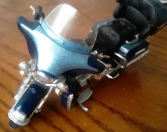 May include: A detailed model motorcycle in shades of blue and silver. The miniature bike features a windshield, saddlebags, and chrome accents. The model is set on a wooden surface.