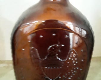 May include: A brown glass bottle with an embossed eagle design. The bottle has a rounded body and a narrow neck. The eagle is centered on the front of the bottle, with its wings spread. The bottle is likely vintage.