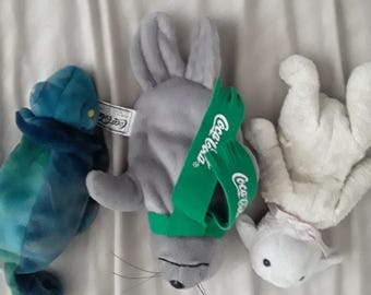 May include: Three plush toys including a blue and green tie-dye chameleon, a gray seal wearing a green scarf with the Coca-Cola logo, and a white lamb with a light pink ribbon around its neck. Each toy has a soft, stuffed fabric texture.