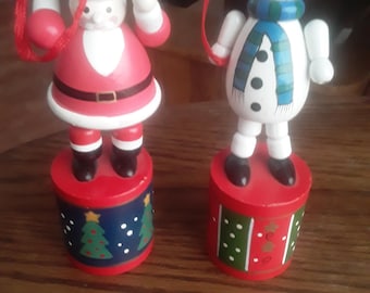 May include: Two wooden Christmas ornaments: Santa Claus and a snowman. The Santa ornament is red and white with a black belt. The snowman is white with a blue and green scarf and a carrot nose. Both ornaments stand on red bases decorated with festive designs.