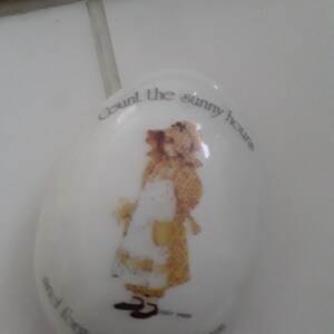 以下が含まれることがあります： 黄色いドレスと白いエプロンを着た女の子が描かれた白い楕円形のセラミック皿。皿の縁には「Count the sunny hours and forget the rainy days」という文字が書かれている。