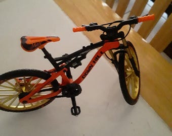 May include: A miniature mountain bike model featuring a bright orange frame with the text DOWN HILL printed on the side. The bicycle has gold-colored wheels, black tires, orange handlebars, and an orange seat with black accents and black pedals.