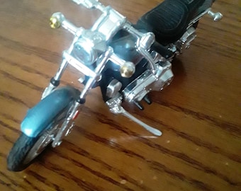 May include: A detailed model motorcycle with a black seat, chrome accents, and a blue front fender. The miniature bike features a black tire and handlebars, and gold-colored lights. The motorcycle is a collectible item.