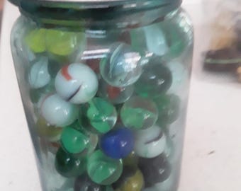 May include: A vintage glass jar filled with colorful marbles. The marbles are various colors, including green, blue, white, and red. The jar has a silver metal lid and a teal-colored glass body. A nostalgic item.
