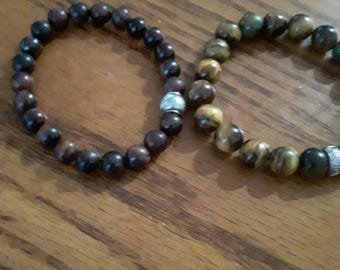 May include: Two beaded bracelets. One bracelet features dark brown beads with a silver accent bead. The other bracelet has brown and gold beads with a silver accent bead. Both bracelets are on a wooden surface.