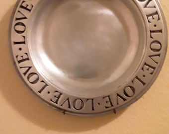 May include: A round, silver-colored metal plate with the word "LOVE" engraved around the rim. The plate is mounted on a light-colored wall. The word "LOVE" is repeated around the edge of the plate.