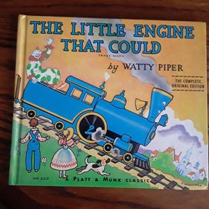 Könnte beinhalten: Ein Vintage-Kinderbuch mit dem Titel "The Little Engine That Could" von Watty Piper. Das Cover zeigt eine blaue Lokomotive, einen Clown und weitere Figuren. Es ist ein Platt & Munk Classic, die vollständige Originalausgabe.