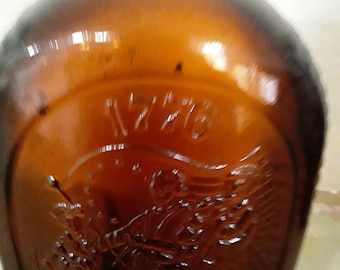 May include: A vintage brown glass bottle with a rounded shoulder and embossed design. The bottle features the year "1776" and an image of a person with wings. The glass has a glossy finish.