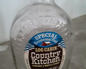May include: Clear glass bottle of Log Cabin Country Kitchen pancake and waffle syrup. The label reads "Special Bicentennial Flask" and "Net 24 fl oz (1 pt 8 oz)". The bottle has a metal cap and is empty.