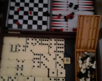 May include: A multi-game set in a wooden case featuring a black and white checkerboard, a backgammon board with red and black points, a collection of off-white dominoes with black pips, and a small plastic container filled with chess pieces.