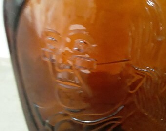 May include: A brown glass bottle with an embossed image of a person wearing glasses. The bottle has a rounded shape and a dark amber color. The embossed image is a detailed depiction of a face.