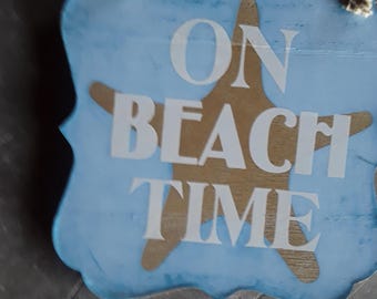May include: A light blue, scalloped-edge wooden wall sign featuring a tan starfish graphic and the white text ON BEACH TIME. The sign is suspended by a natural jute rope attached to two metal eyelets at the top of the wooden plaque.