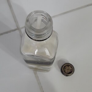 May include: A clear, square glass bottle with a silver and black band around the middle. The bottle is partially filled with a white substance. A small, round cap with a gold design sits next to the bottle.