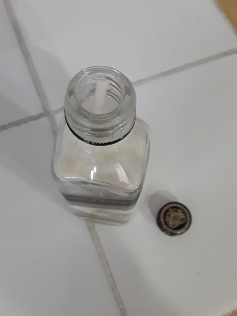 May include: A clear, square glass bottle with a silver and black band around the middle. The bottle is partially filled with a white substance. A small, round cap with a gold design sits next to the bottle.