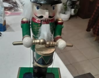 May include: A wooden nutcracker figurine in a green and red uniform, holding drumsticks and a drum. The nutcracker has a white beard, a black hat with gold accents, and a stern expression. The base is green with gold trim.