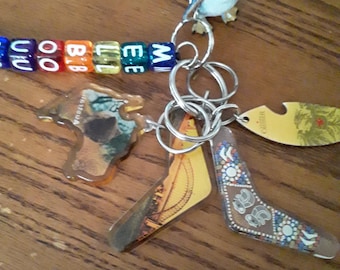 May include: A collection of keychains featuring a blue bird figurine, colorful letter beads spelling out a word, and several boomerang-shaped charms. The charms display various designs and images, including a landscape and an abstract pattern.