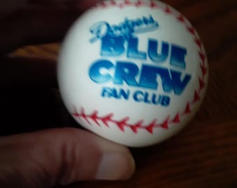 May include: A white baseball with red stitching and the text "Dodgers BLUE CREW FAN CLUB" in blue. The baseball is held in a person's hand.