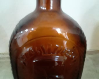 May include: A brown glass bottle with a rounded body and a narrow neck. The bottle has a decorative embossed design on the front and a cork stopper. The glass has a glossy finish.
