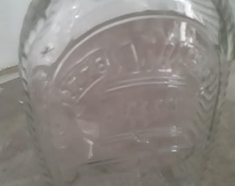 May include: Clear glass bottle with a rounded body and a narrow neck. The bottle features embossed details, including a crown and text within a circular design. The glass has a slightly textured appearance.