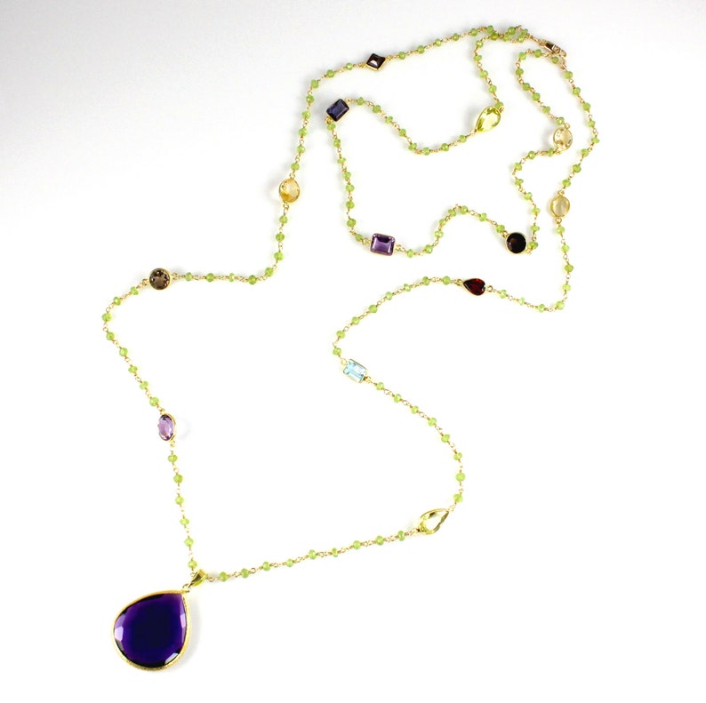 May include: A long gold chain necklace with a purple teardrop pendant. The chain is made of small green beads and features various coloured gemstones.