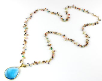 May include: A long gold chain necklace with a teardrop-shaped blue gemstone pendant. The chain is adorned with small, colorful beads.