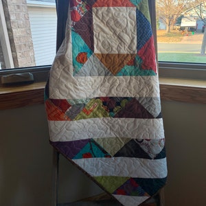 May include: A colorful patchwork quilt with a white background and a brown, orange, teal, and purple geometric pattern. The quilt is folded and draped over a wooden ladder.
