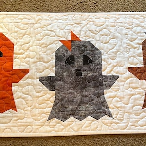 May include: A rectangular quilted table runner featuring a white background with a swirling stitch pattern. Three ghost-shaped fabric appliqués are centered in a row, colored orange, dark gray, and brown, each with black eyes and a mouth.