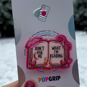 Puede incluir: Agarre para teléfono rosa y blanco con un diseño de libro. El libro tiene el texto "Don't Ask Me" en un lado y "What I'm Reading" en el otro. El agarre para teléfono es de la marca PopGrip.