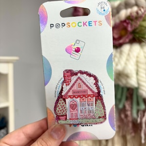 Puede incluir: Un agarre para teléfono rosa y blanco con un diseño de casa de jengibre. La casa tiene una puerta y ventanas rojas, y el texto "BOOKSTORE" está escrito sobre la puerta. El agarre está diseñado para ser usado con una funda de teléfono.