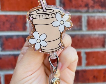 May include: A gold glitter badge reel with a clear plastic holder. The design features a cartoon illustration of a brown iced coffee cup with a straw and pink flowers.