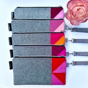 May include: A stack of five rectangular pencil cases. The cases are gray with a color block design in shades of pink, red, and orange. Each case has a zipper closure and a wrist strap. The cases are arranged on a white surface.
