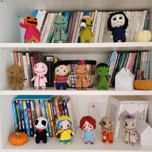 May include: A collection of handmade crochet dolls, each representing a different character. The dolls are displayed on a white shelf in front of a backdrop of books. The dolls include a pumpkin, zombie, and other spooky characters.
