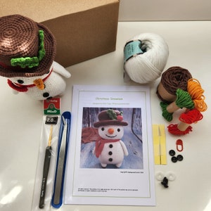 May include: A crochet Christmas snowman kit with white, brown, red, green, and orange yarn, a crochet hook, a needle, buttons, and a pattern. The pattern is titled "Christmas Snowman" and is designed by "Heidi Tagle @WhimsygnomeCrochet".