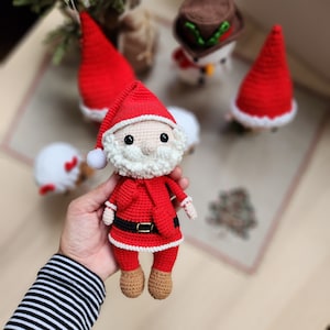 May include: A crocheted Santa Claus doll with a white beard and red and white hat. The doll is wearing a red and white suit with a black belt and is holding a red bag.