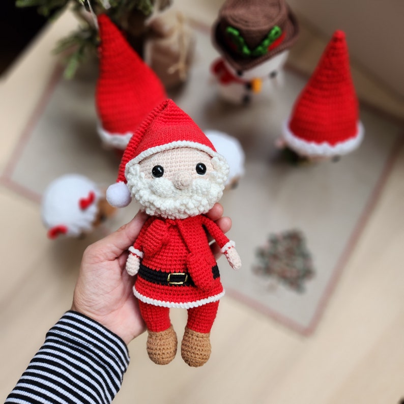 May include: A crocheted Santa Claus doll with a white beard and red and white hat. The doll is wearing a red and white suit with a black belt and is holding a red bag.